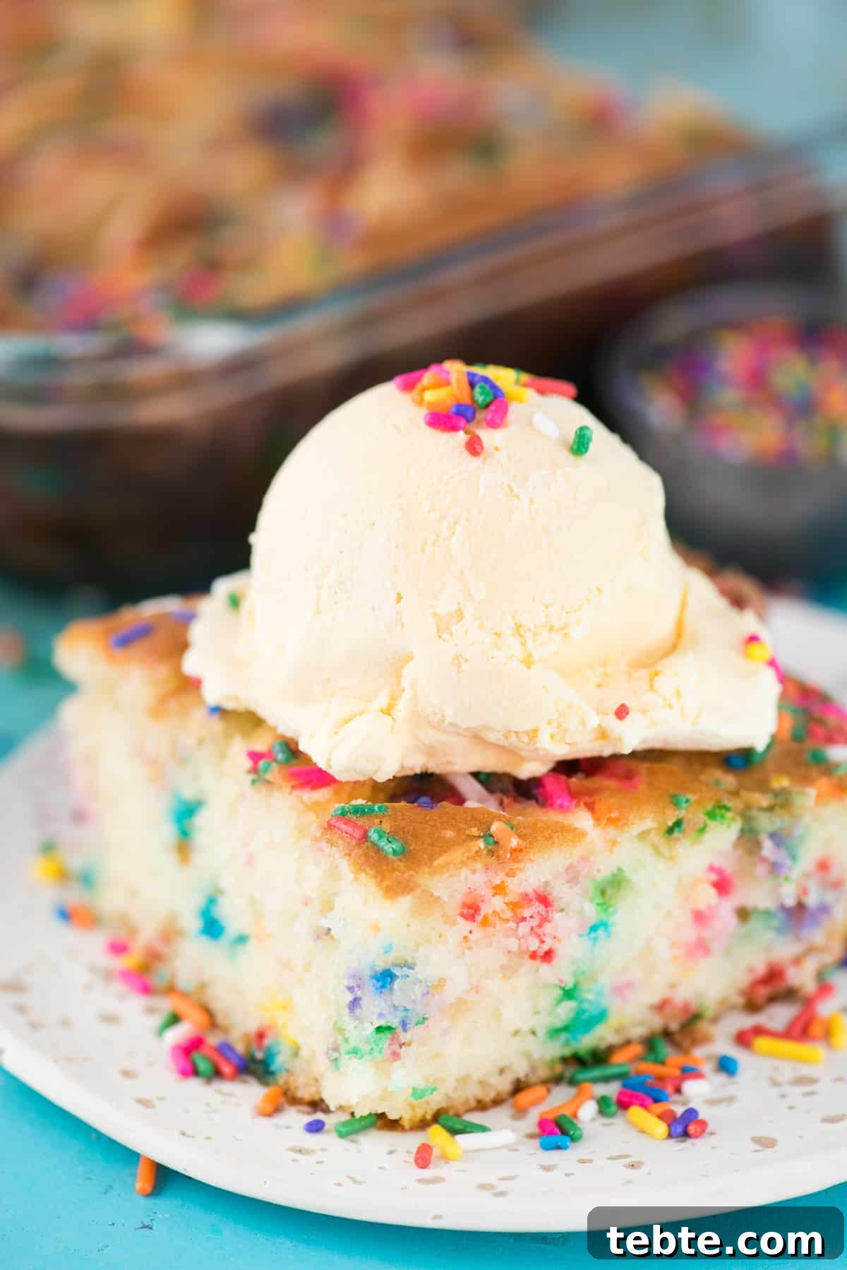 This funfetti earthquake cake is a white cake mix loaded with sprinkles and funfetti chocolate chips. Swirl in the cream cheese frosting layer and it bakes into the cake creating crevices and cracks on top of the cake. #earthquakecake #funfetticake #cake slice of funfetti earthquake cake with scoop of vanilla ice cream on white plate on blue background