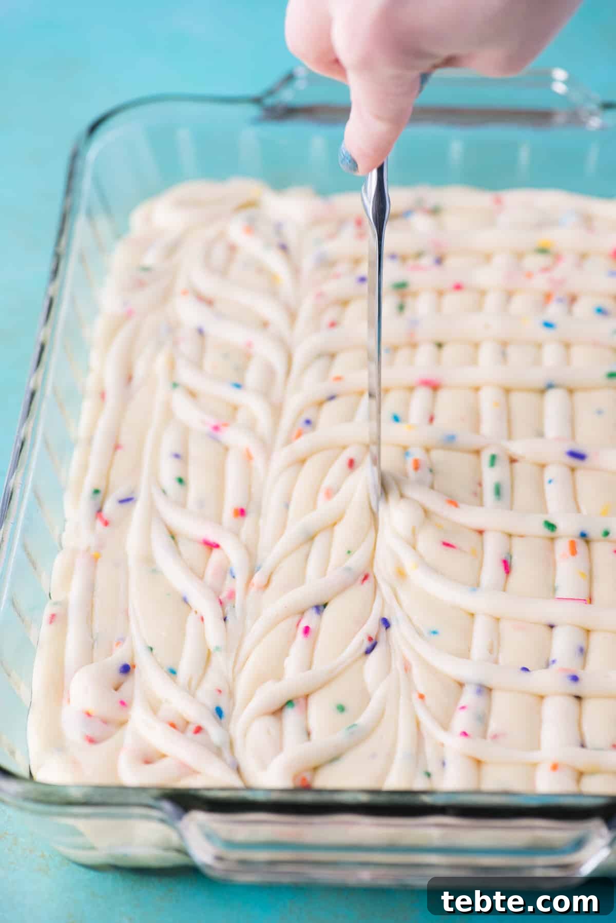 This funfetti earthquake cake is a white cake mix loaded with sprinkles and funfetti chocolate chips. Swirl in the cream cheese frosting layer and it bakes into the cake creating crevices and cracks on top of the cake. #earthquakecake #funfetticake #cake swirling cake batter and cream cheese frosting together with butter knife in glass 9x13 inch pan