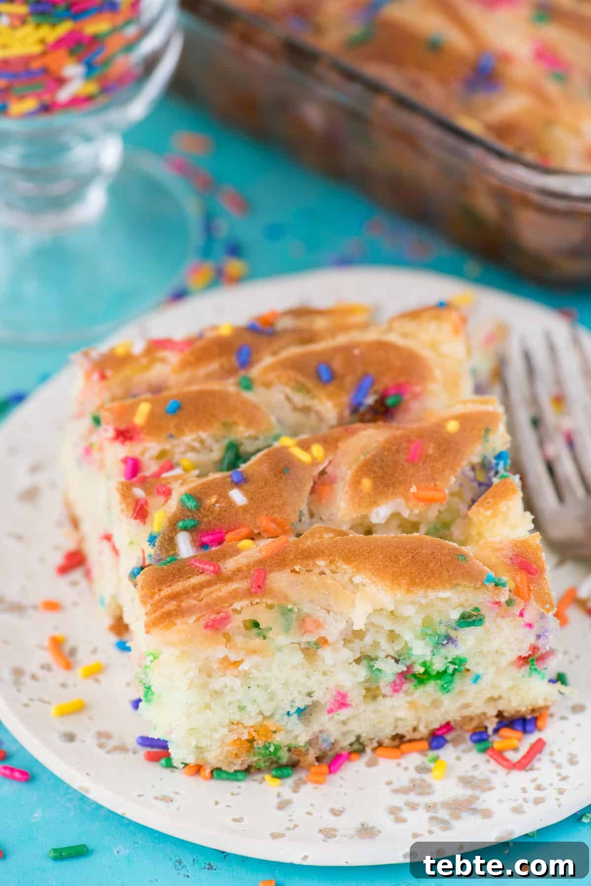 This funfetti earthquake cake is a white cake mix loaded with sprinkles and funfetti chocolate chips. Swirl in the cream cheese frosting layer and it bakes into the cake creating crevices and cracks on top of the cake. #earthquakecake #funfetticake #cake slice of funfetti earthquake cake on white plate on blue background