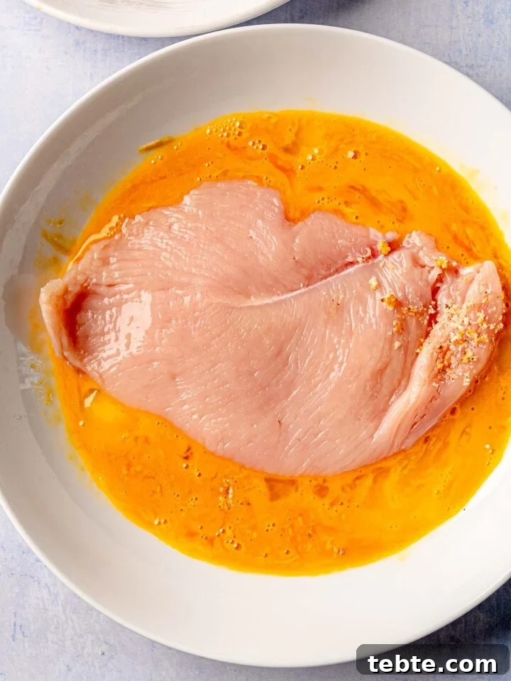 A raw chicken breast coated in an egg and mustard mixture, resting in a white plate, ready to be breaded.