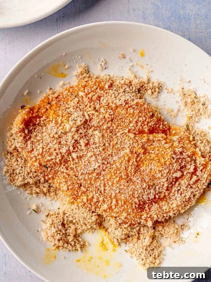 A chicken breast coated in a seasoned panko and parmesan breadcrumb mixture, sitting on a white plate.