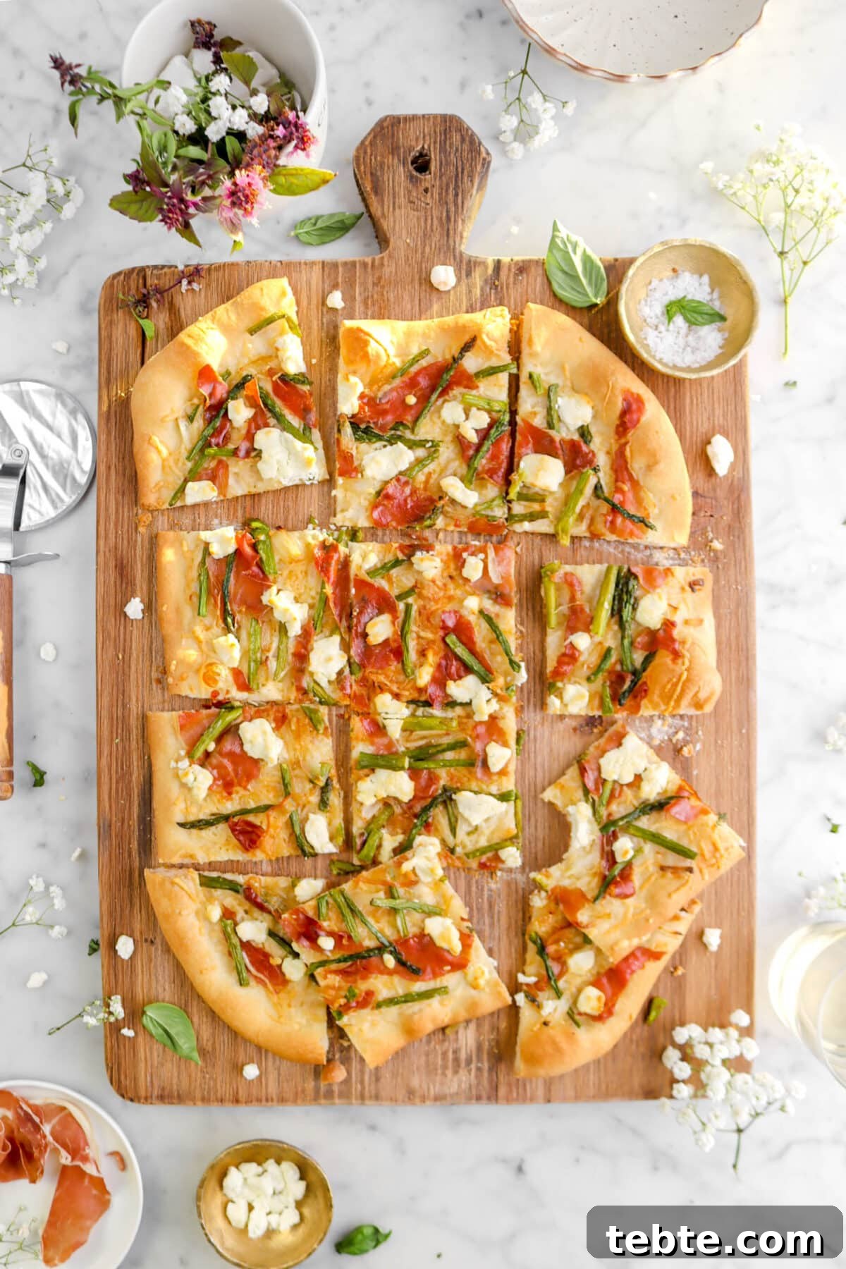 Asparagus, Caramelized Onion & Prosciutto Flatbread with Goat Cheese 19 sliced asparagus and prosciutto flatbread on wood board with one slice missing a bite, a gold bowl beside, fresh flowers, and basil leaves around on marble surface.