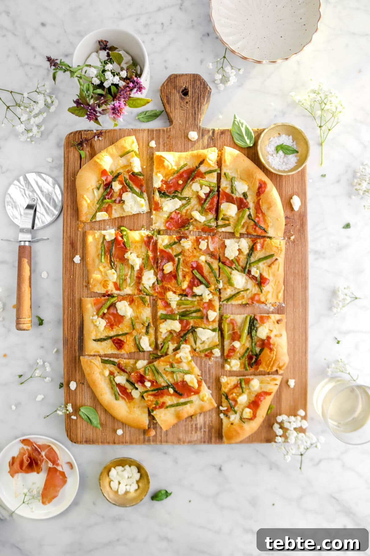 Asparagus, Caramelized Onion & Prosciutto Flatbread with Goat Cheese 4 sliced asparagus and prosciutto flatbread on wood board with basil leaves and flowers around on marble surface.