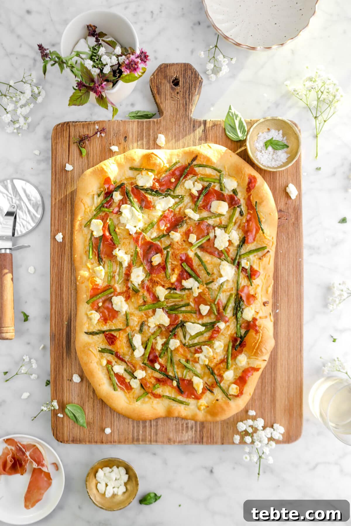 Asparagus, Caramelized Onion & Prosciutto Flatbread with Goat Cheese 6 flatbread on wood board with fresh herbs and flowers around with sunlight streaming through.