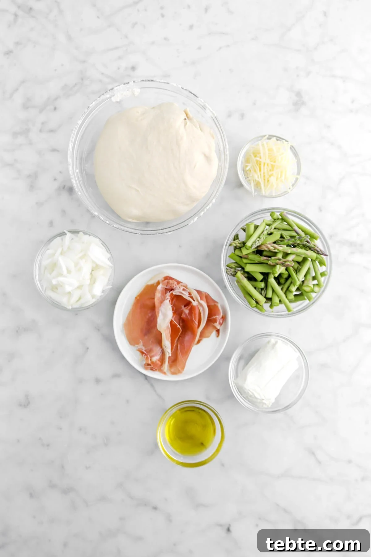 Asparagus, Caramelized Onion & Prosciutto Flatbread with Goat Cheese 7 pizza dough, grated parmesan cheese, slices onion, prosciutto, asparagus tops, log of goat cheese, and olive oil on marble surface.