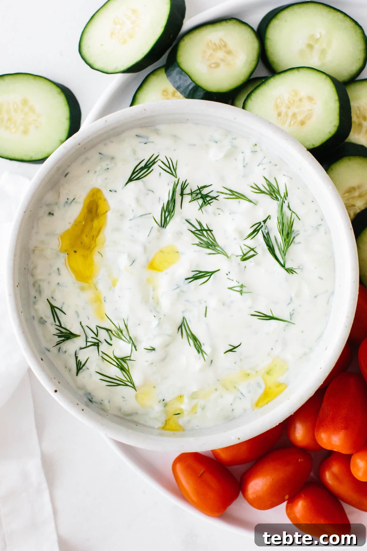 Tzatziki in a bowl surrounded by sliced cucumber and cherry tomatoes.
