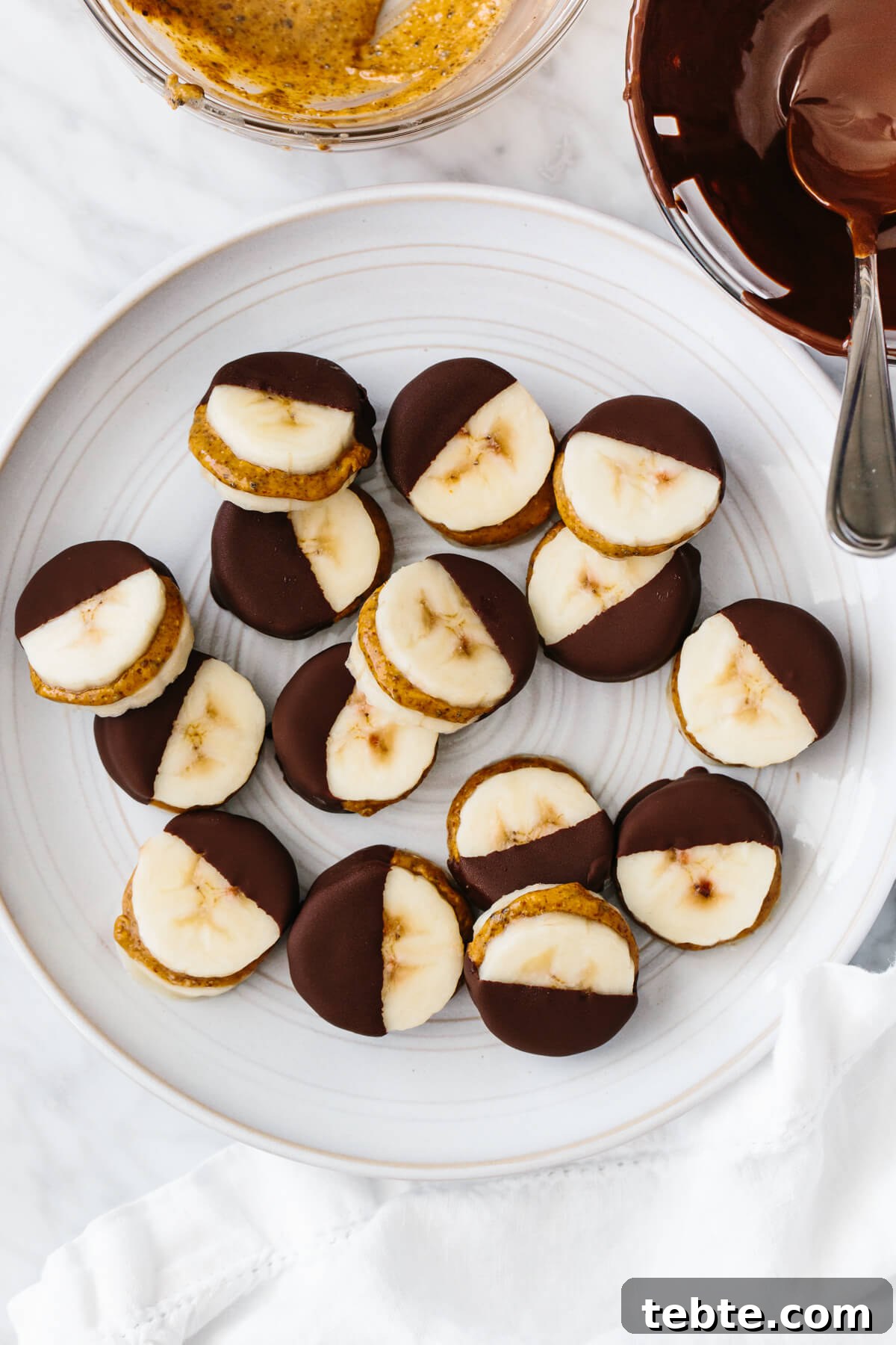 Frozen chocolate banana bites on a plate next to bowls of chocolate and almond butter.