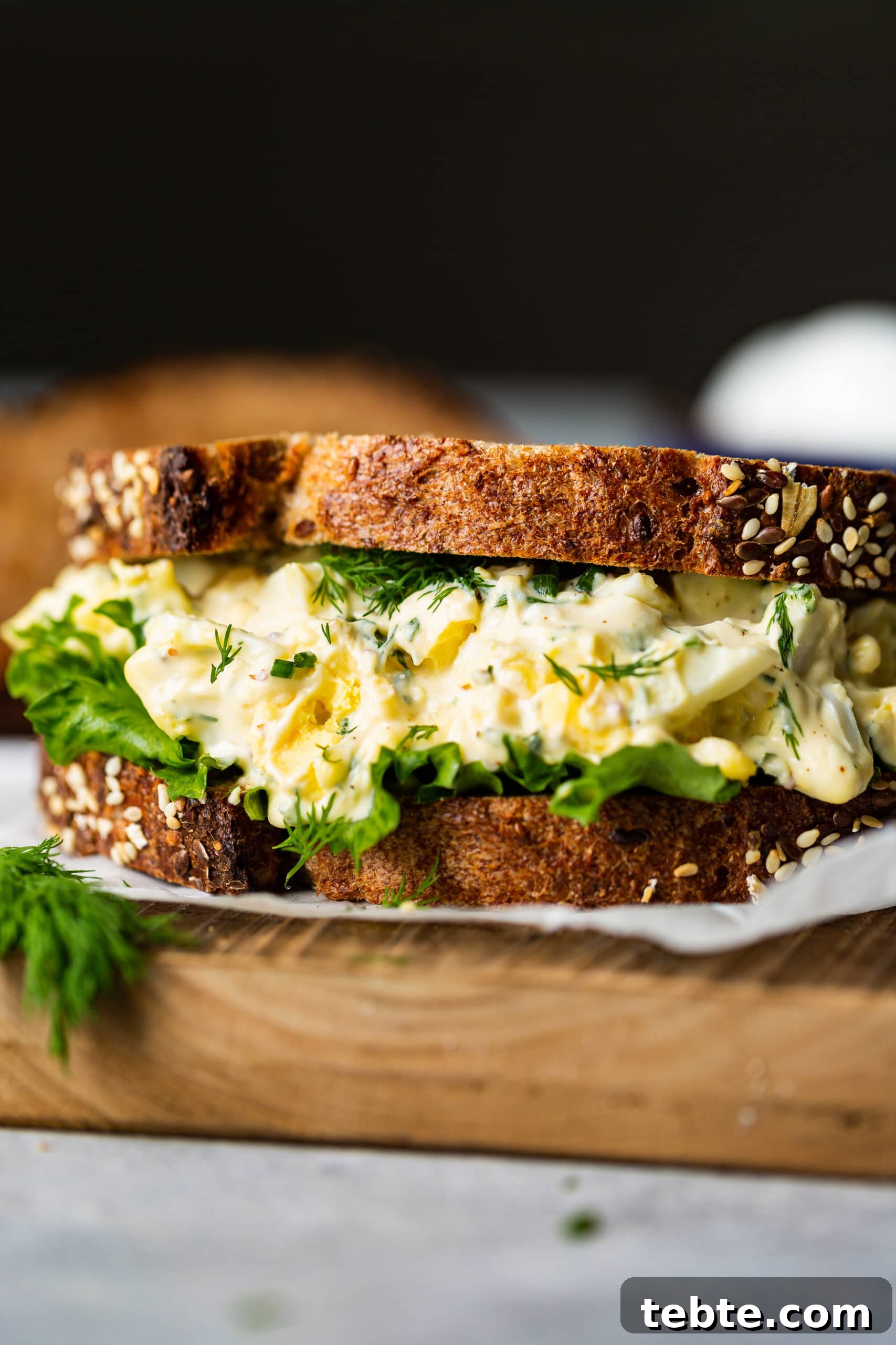 The BEST Egg Salad Sandwich Recipe a photo of an egg salad sitting on a piece of parchment paper made on multigrain bread with lettuce and thick layer of egg salad.