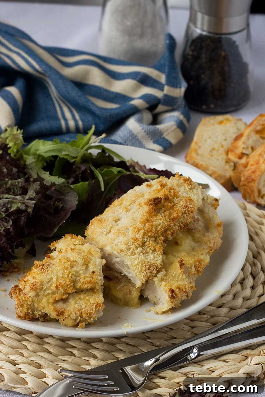 Crispy Baked Chicken Stuffed with Ham and Melted Cheese 3 Crispy baked cheese and ham stuffed chicken on a white plate with salad and with a blue tea towel and bread in the background.
