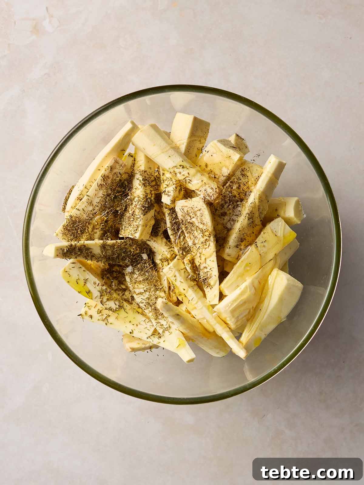 Crispy Roasted Parsnips with Honey and Rosemary 6 A glass bowl on a white surface, filled with raw chopped parsnips that's covered with dried thyme and honey.