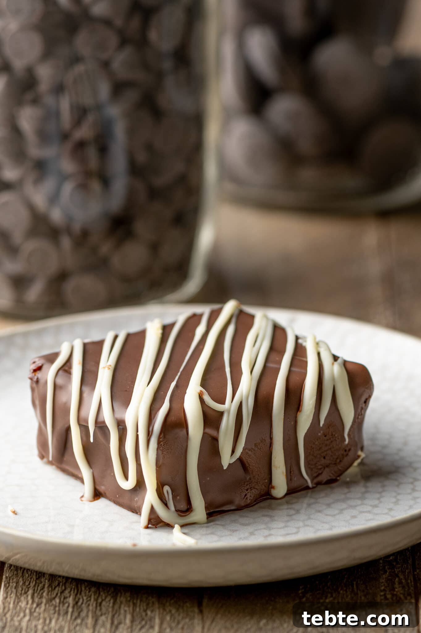 A chocolate covered square of cheesecake on a white texture plate against a rustic brown background.
