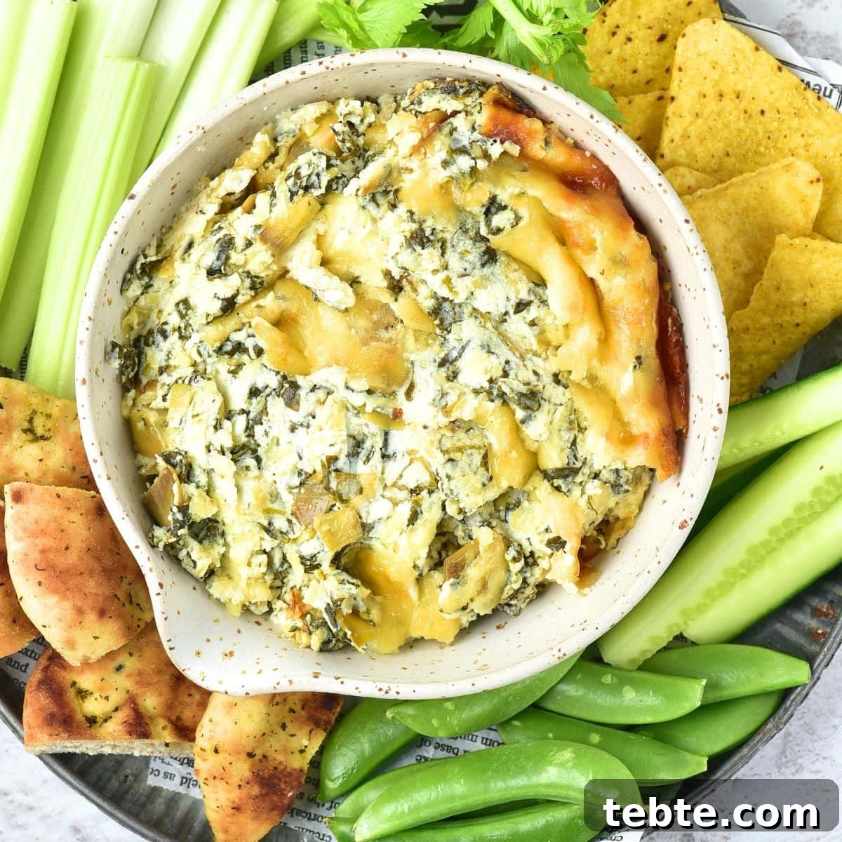 Top down view of a bowl of spinach artichoke dip surrounded with vegetable crudités and chips.
