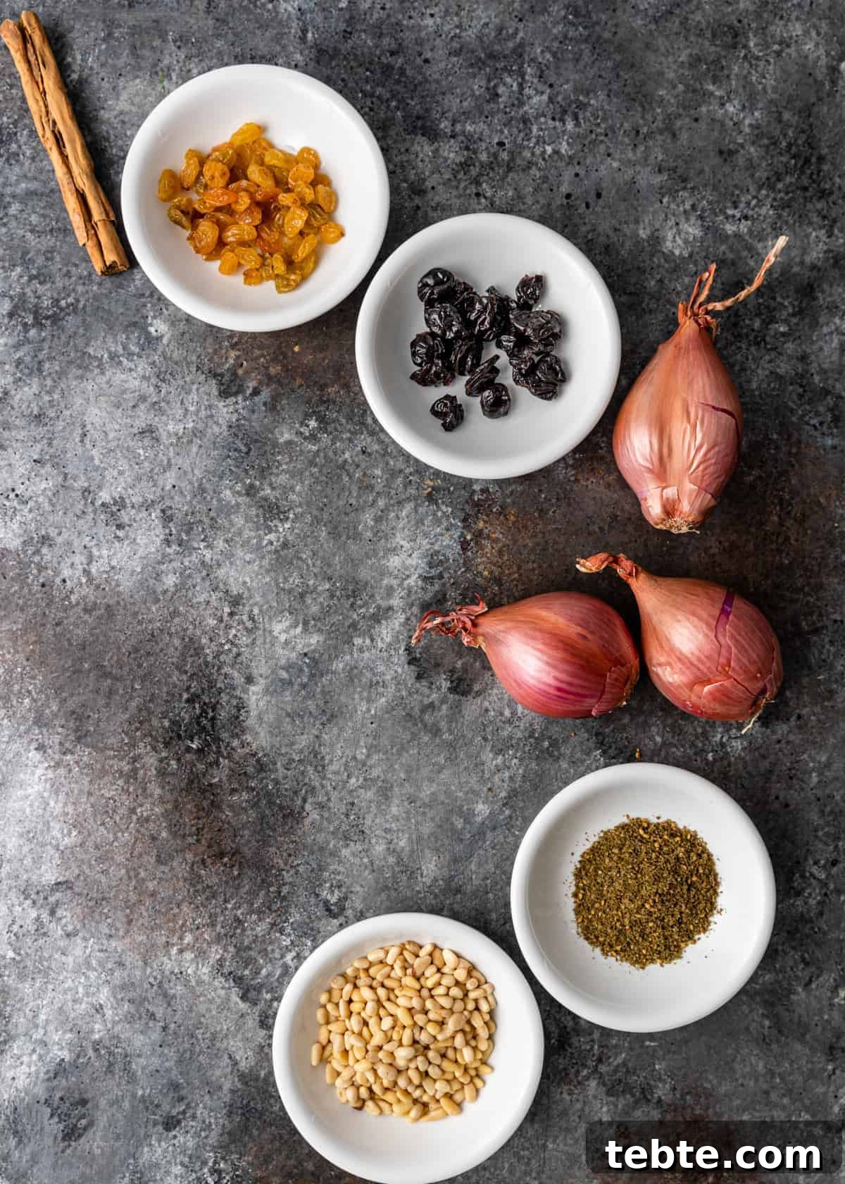 Authentic Middle Eastern Rice Pilaf Recipe with Aromatic Spices 4 overhead: bowls of golden raisins, dried cherries, za'atar spice and pine nuts next to whole, 3unpeele d shallots