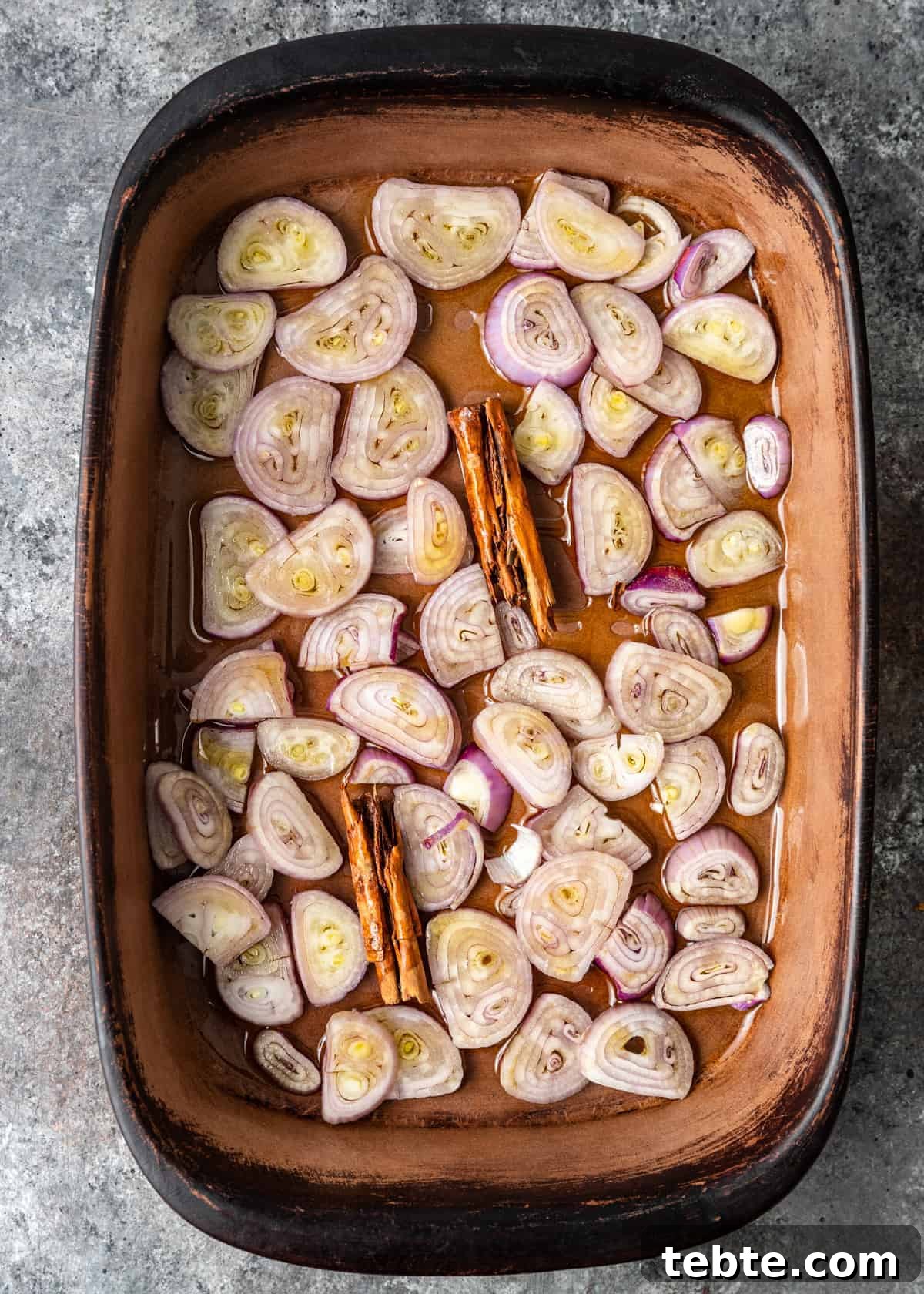 Authentic Middle Eastern Rice Pilaf Recipe with Aromatic Spices 5 overhead: thin slices of shallot in baking dish with cinnamon sticks, ready for oven roasting