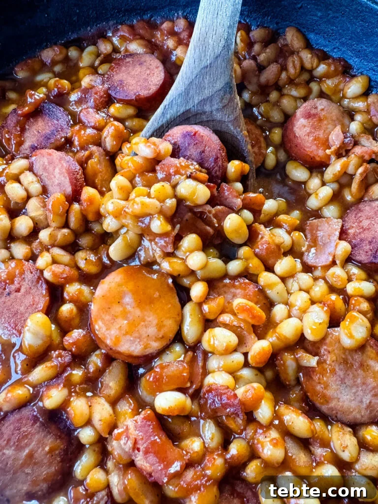 Classic Homemade Pork and Beans Recipe for Comforting Meals 11 homemade pork and beans with sausage, bacon, and a wooden spoon in a cast iron skillet
