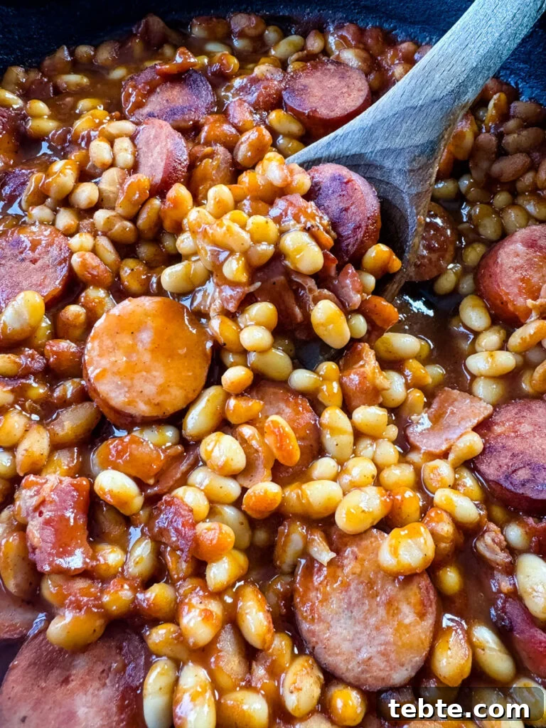 Classic Homemade Pork and Beans Recipe for Comforting Meals 9 homemade pork and beans with sausage, bacon, and a wooden spoon in a cast iron skillet