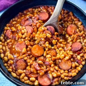 Classic Homemade Pork and Beans Recipe for Comforting Meals 10 homemade pork and beans with sausage, bacon, and a wooden spoon in a cast iron skillet
