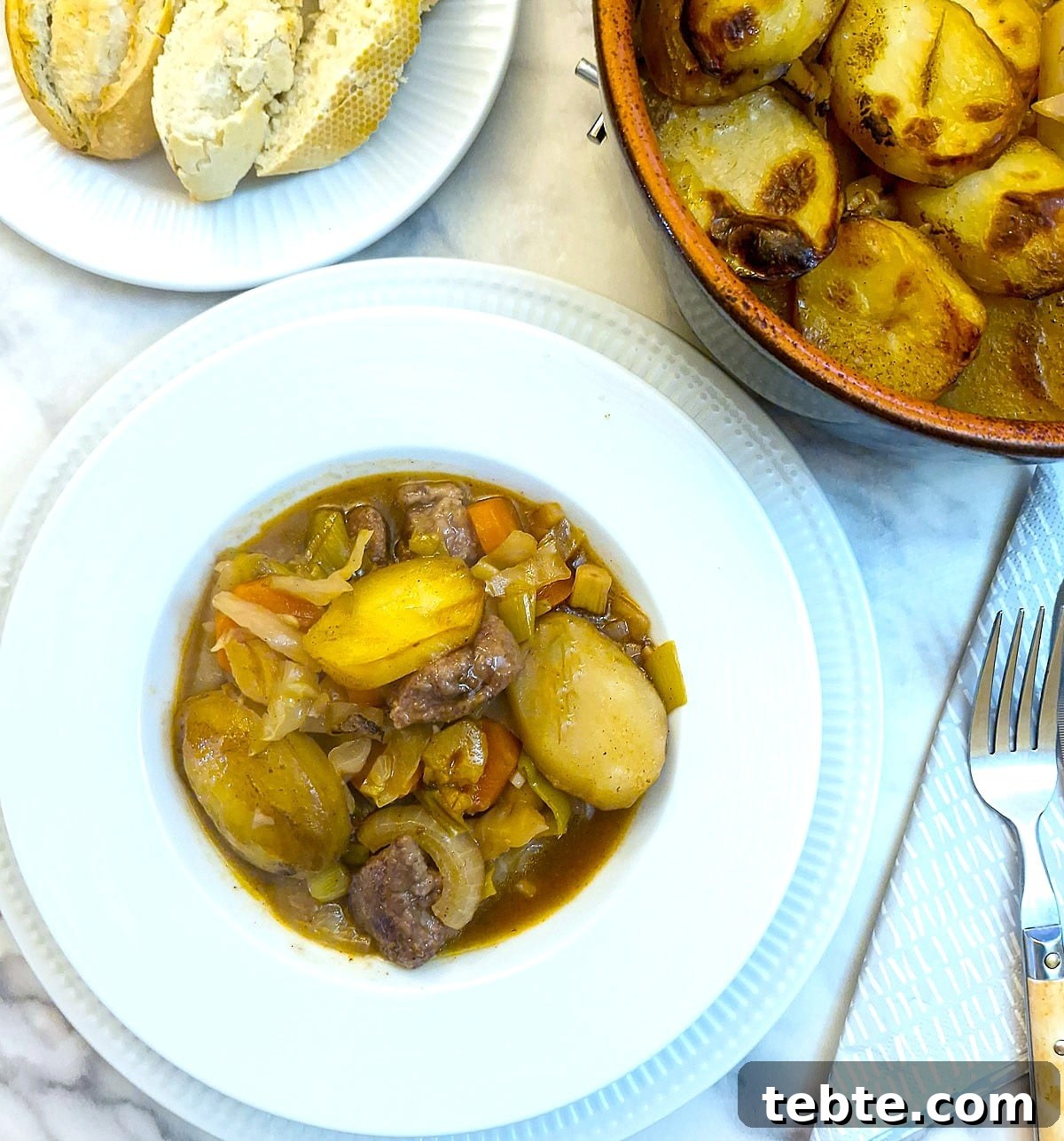 A white dinner plate with a helping of lamb potjie. There is a plate of crusty bread and the serving dish of lamb stew alongside on the table.