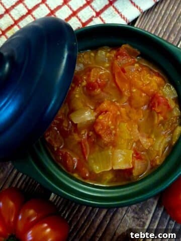 A green serving dish filled with delicious tomato and onion stew. The lid of the dish is propped on the side.