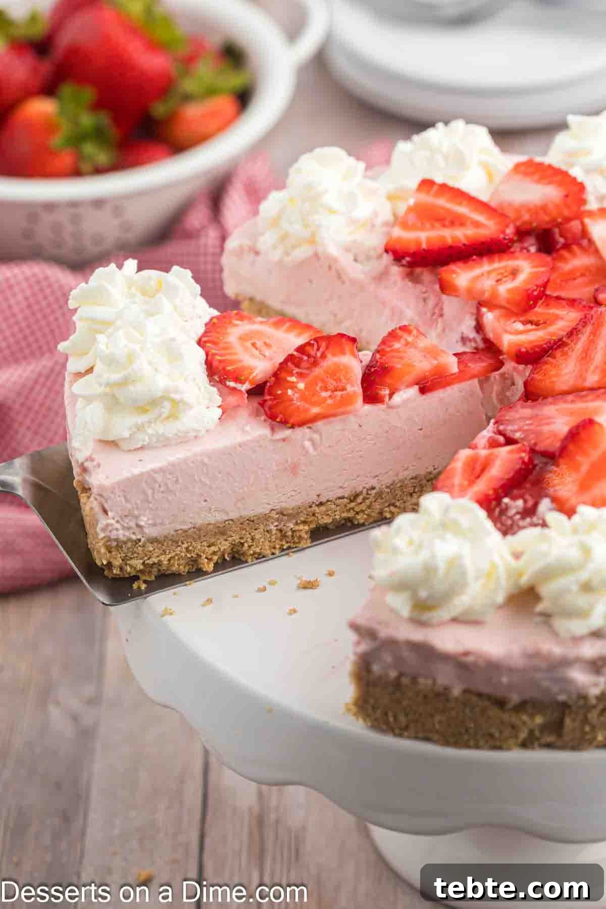 Creamy No-Bake Strawberry Cheesecake Recipe 2 Strawberry Cheesecake topped with sliced strawberries and whipped cream on a cake stand. A slice on a spatula ready to serve.