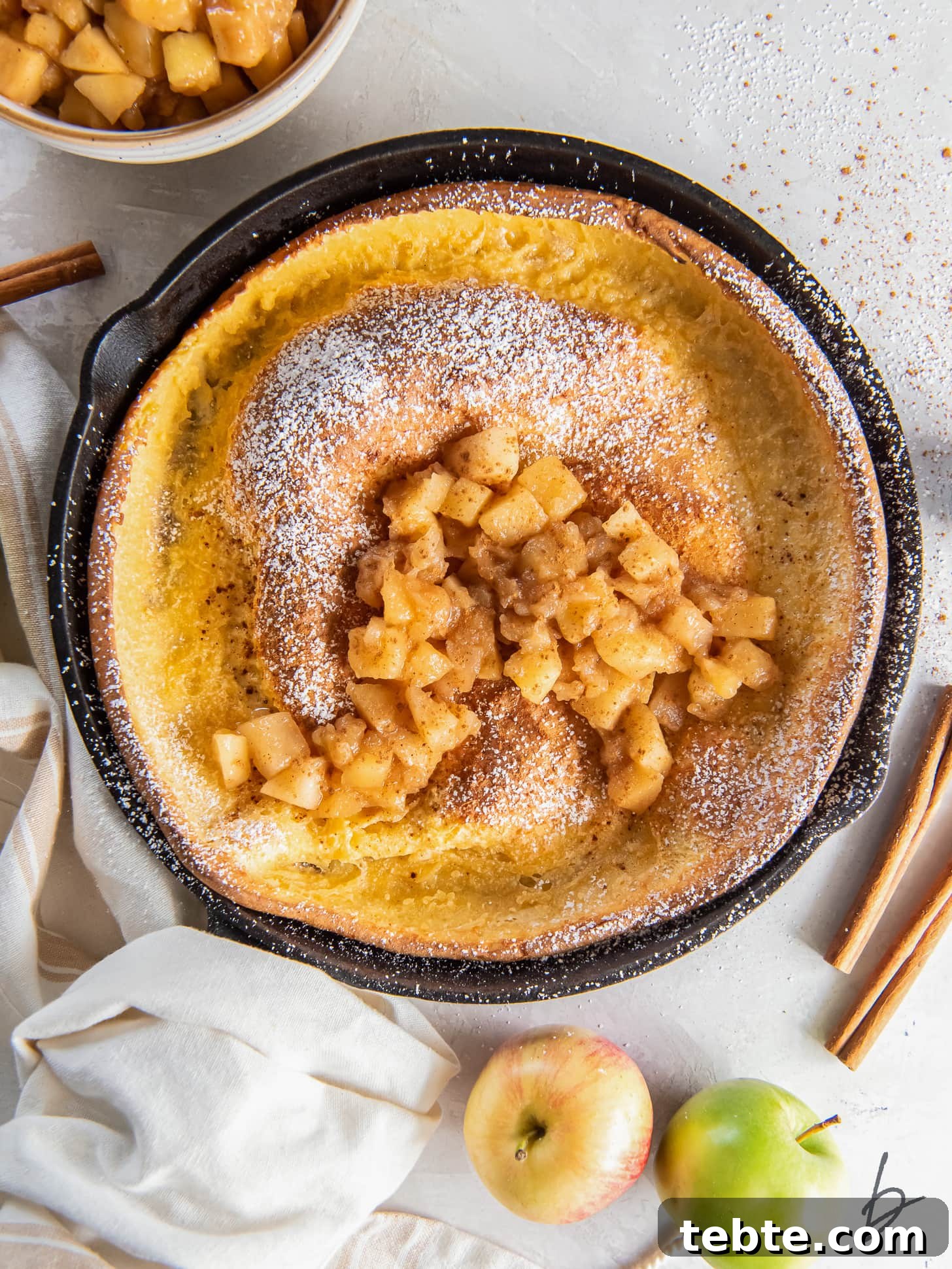 Classic Apple Dutch Baby Pancake Recipe for Brunch 2 apple dutch baby in a cast iron skillet with hand wrapped with kitchen towel.