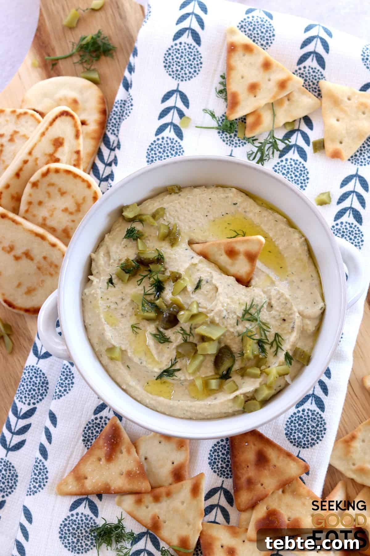 Dill Pickle Hummus: Tangy Chickpea Spread Recipe That Pops 3 Creamy dill pickle hummus served in a white round crock and garnished with chopped dill pickles and served with pita chips.