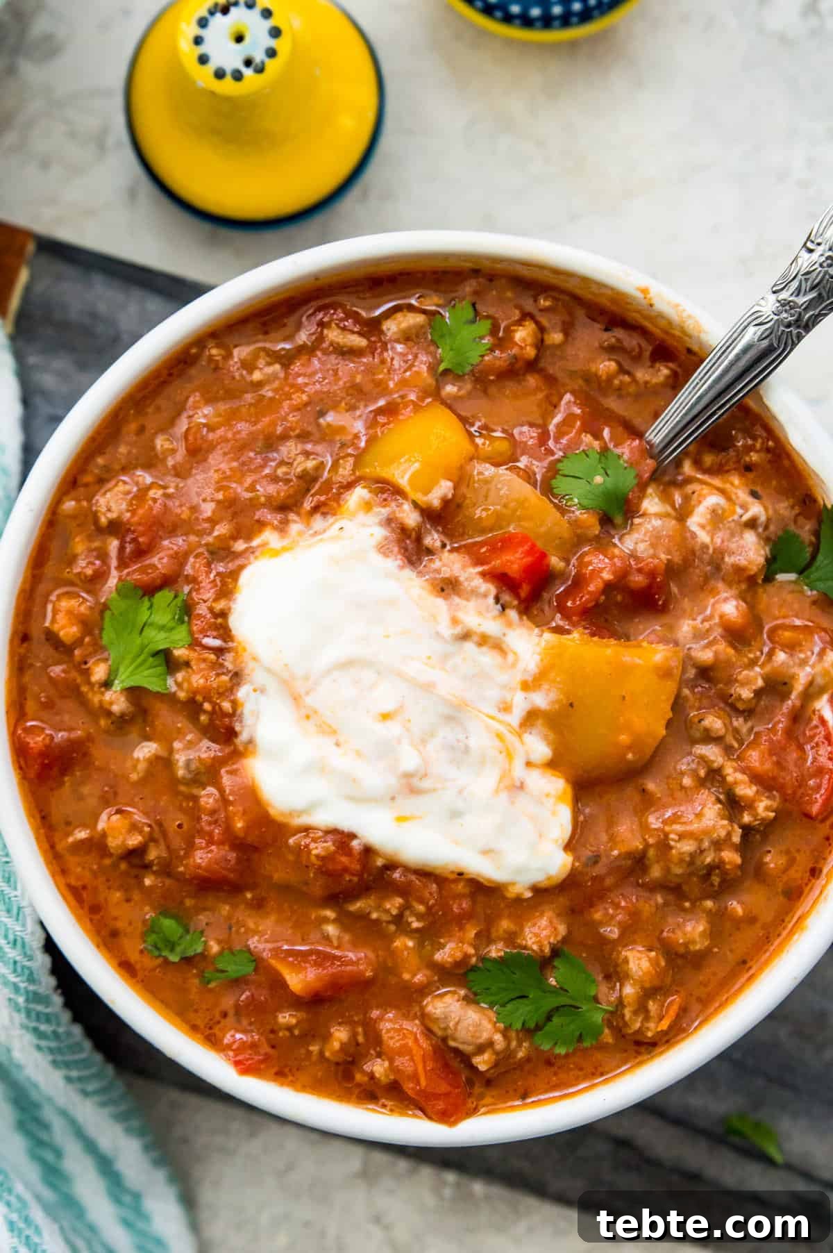 A bowl of ground pork chili topped with sour cream with a spoon in it and garnished with fresh cilantro. 
