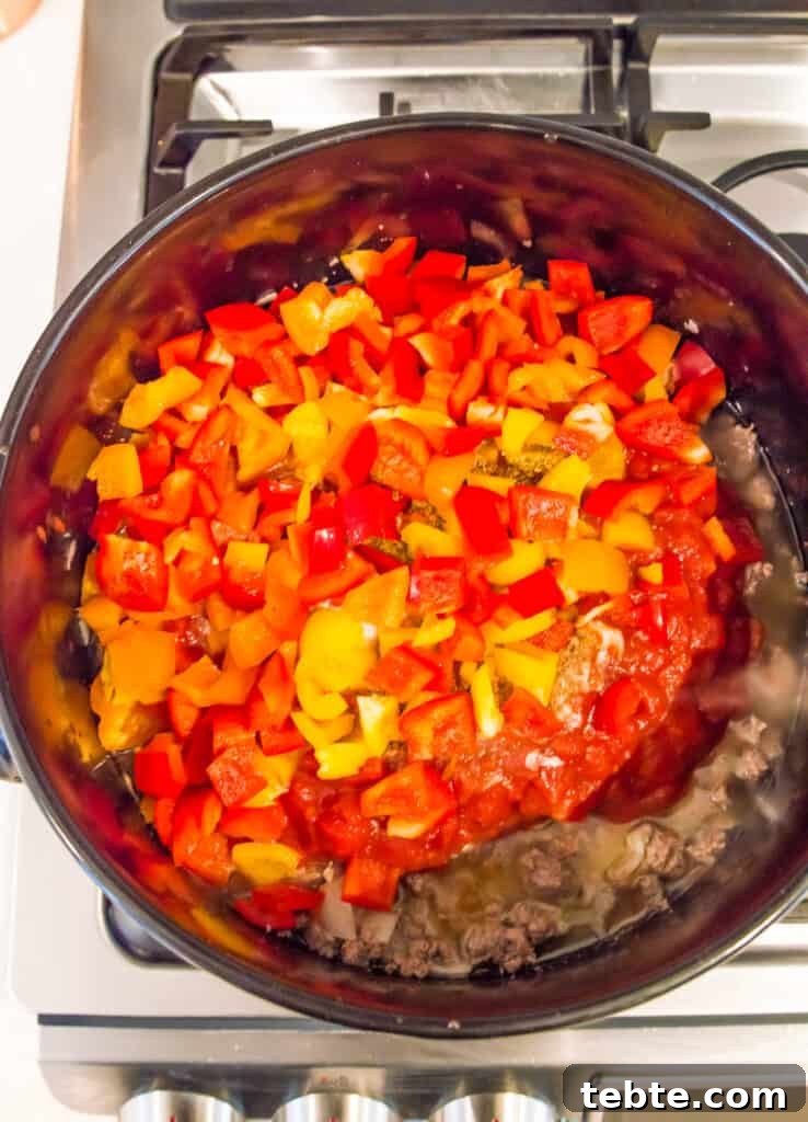 A pot on the stovetop with cooked ground meat, tomatoes and chopped bell peppers in it.
