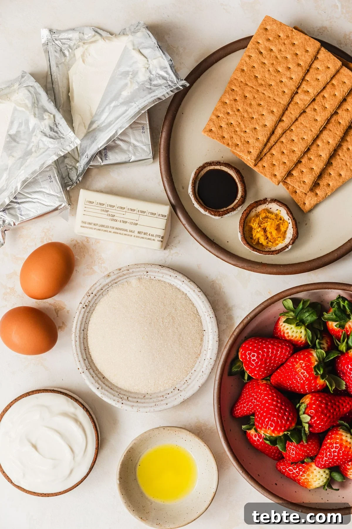 Mini Strawberry Cheesecakes Recipe: Perfect Bite-Sized Treats 3 White and brown bowls of sugar, graham crackers, vanilla, lemon, strawberries, limoncello, and sour cream on a tan counter next to a stick of butter, eggs, and two blocks of cream cheese.