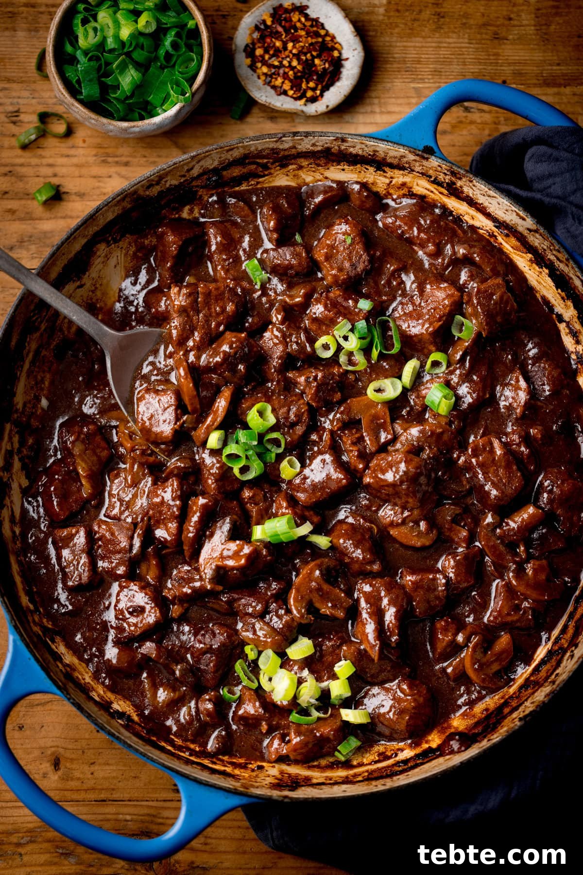 Chinese-Style Beef Curry Recipe: Savory Stir-Fry with Aromatics 2 A blue cast iron casserole filled with Chinese Beef Curry, topped with sliced spring onions. There are little bowls of chopped spring onions and chilli flakes nearby. The pan is on a wooden surface.