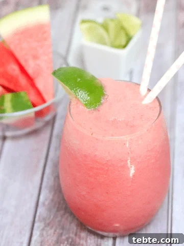 Pink watermelon slushie in a glass garnished with lime and more watermelon and lime behind it