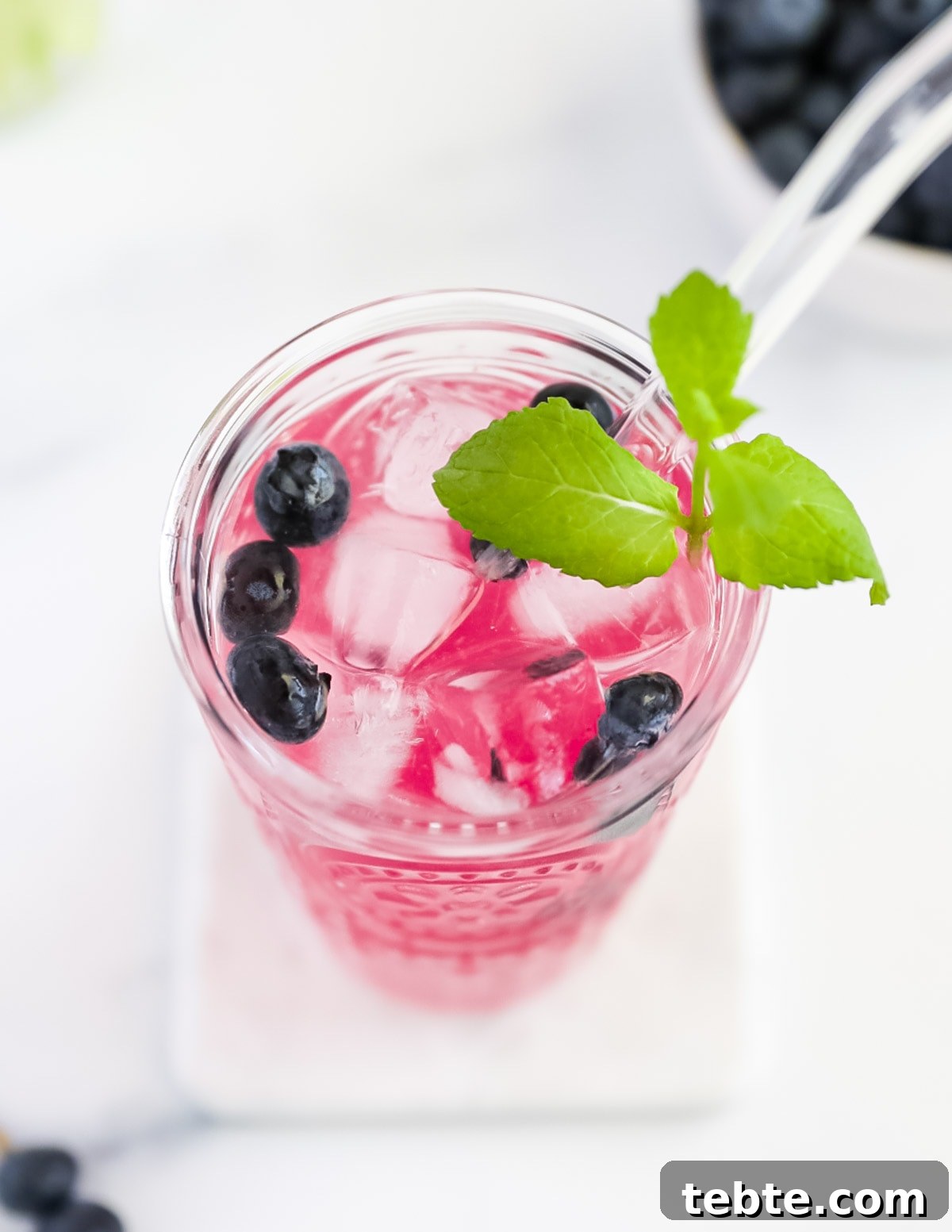 An overhead picture of a glass filled with bright pink lemonade. There are berries, mint, and ice cubes, along with a glass straw in the cup.