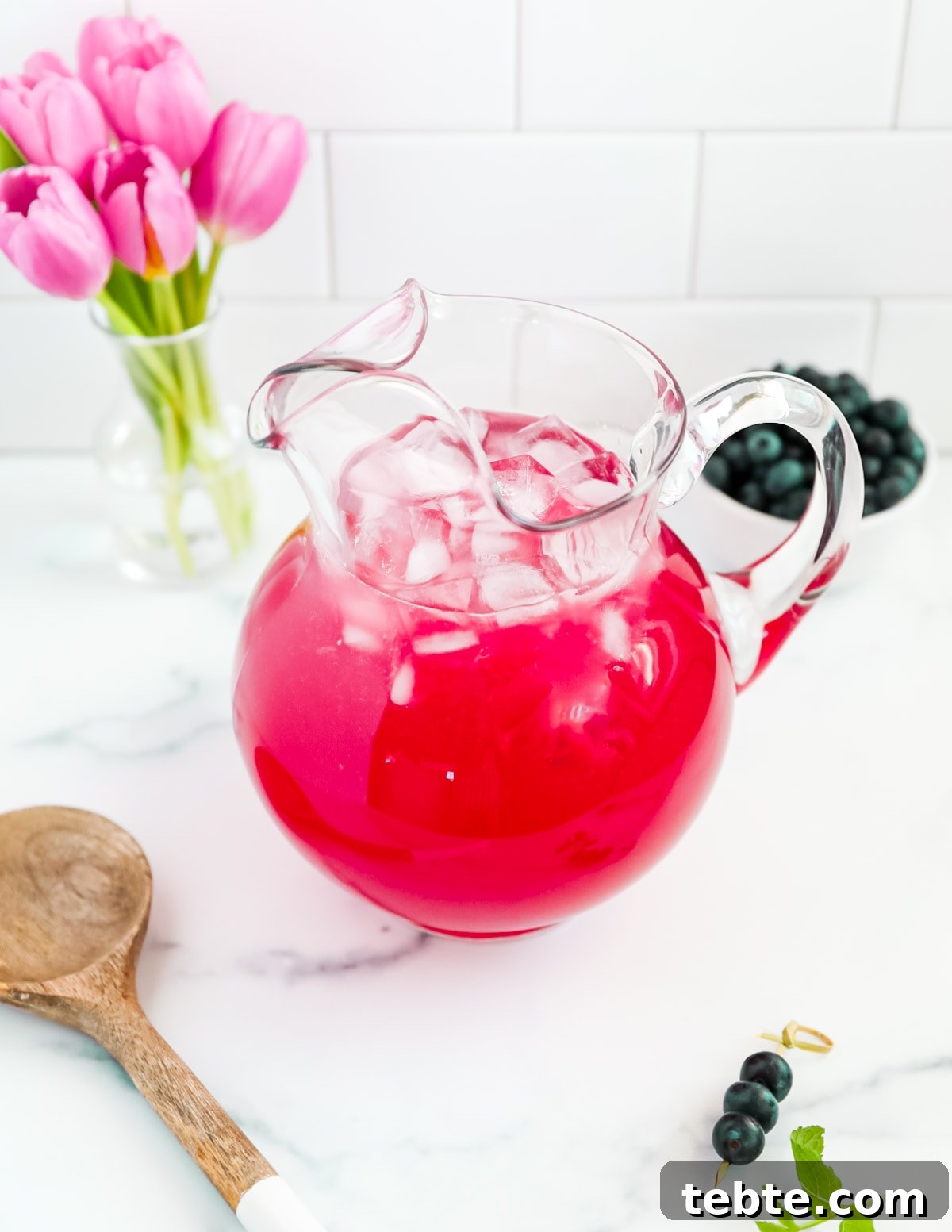 A clear glass pitcher filled with bright pink lemonade. There are ice cubes in the pitcher and flowers, berries, and a spoon outside