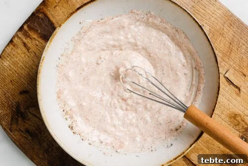 Creamy Yogurt-Marinated Chicken Breast Recipe 10 A bowl of creamy, light pink sauce—perfect for Yogurt Marinated Chicken Breast—speckled with seasoning is whisked with a metal whisk with a wooden handle, set on a wooden surface.