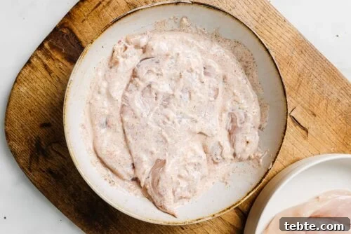 Creamy Yogurt-Marinated Chicken Breast Recipe 11 Yogurt Marinated Chicken Breast pieces coated in a creamy, seasoned marinade sit in a shallow white bowl on a wooden cutting board, with a plate of plain chicken breasts nearby.