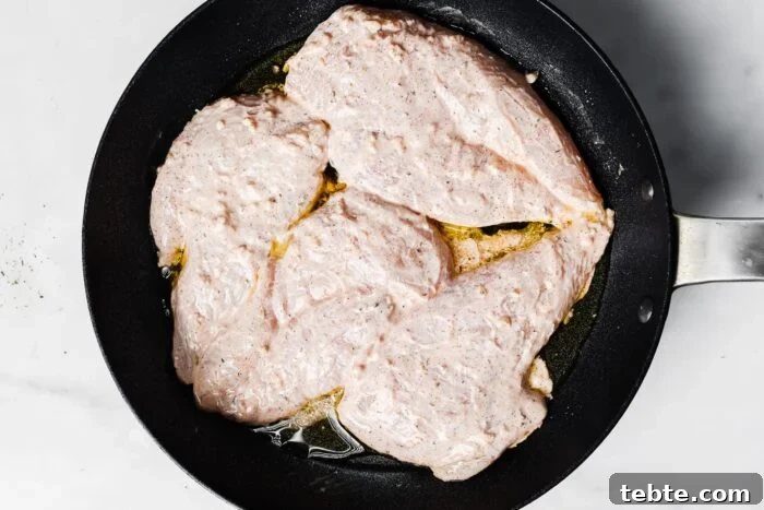 Creamy Yogurt-Marinated Chicken Breast Recipe 6 Raw, seasoned Yogurt Marinated Chicken Breasts are coated in a creamy marinade and spread out in a black frying pan with some oil, ready to be cooked, photographed from above on a white surface.