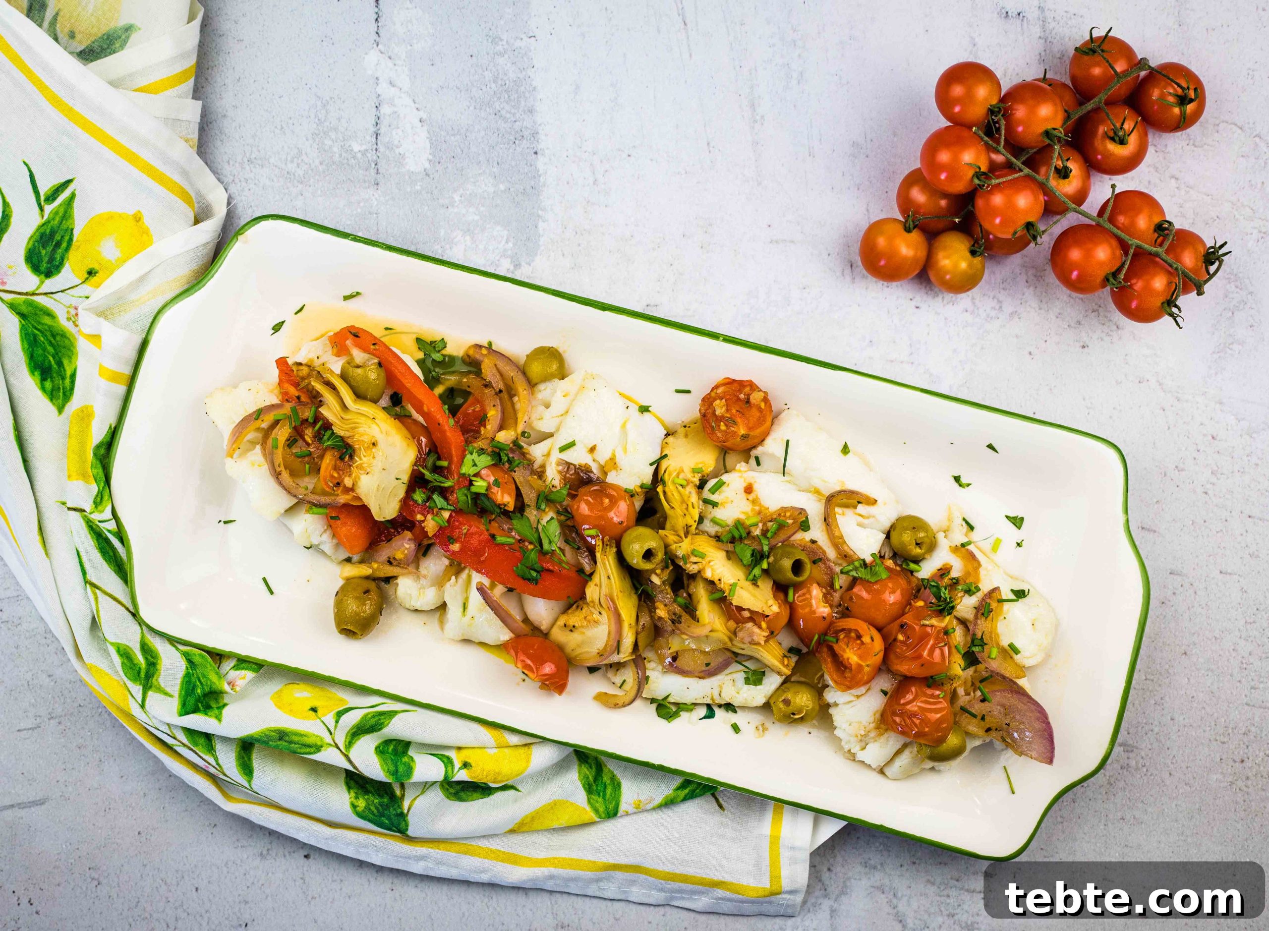 15 Easter Recipes Your Family Will Beg to Eat This Year 5 A rectangular plate with Mediterranean Cod placed on a lemon-patterned cloth. A vine of cherry tomatoes is beside the plate.