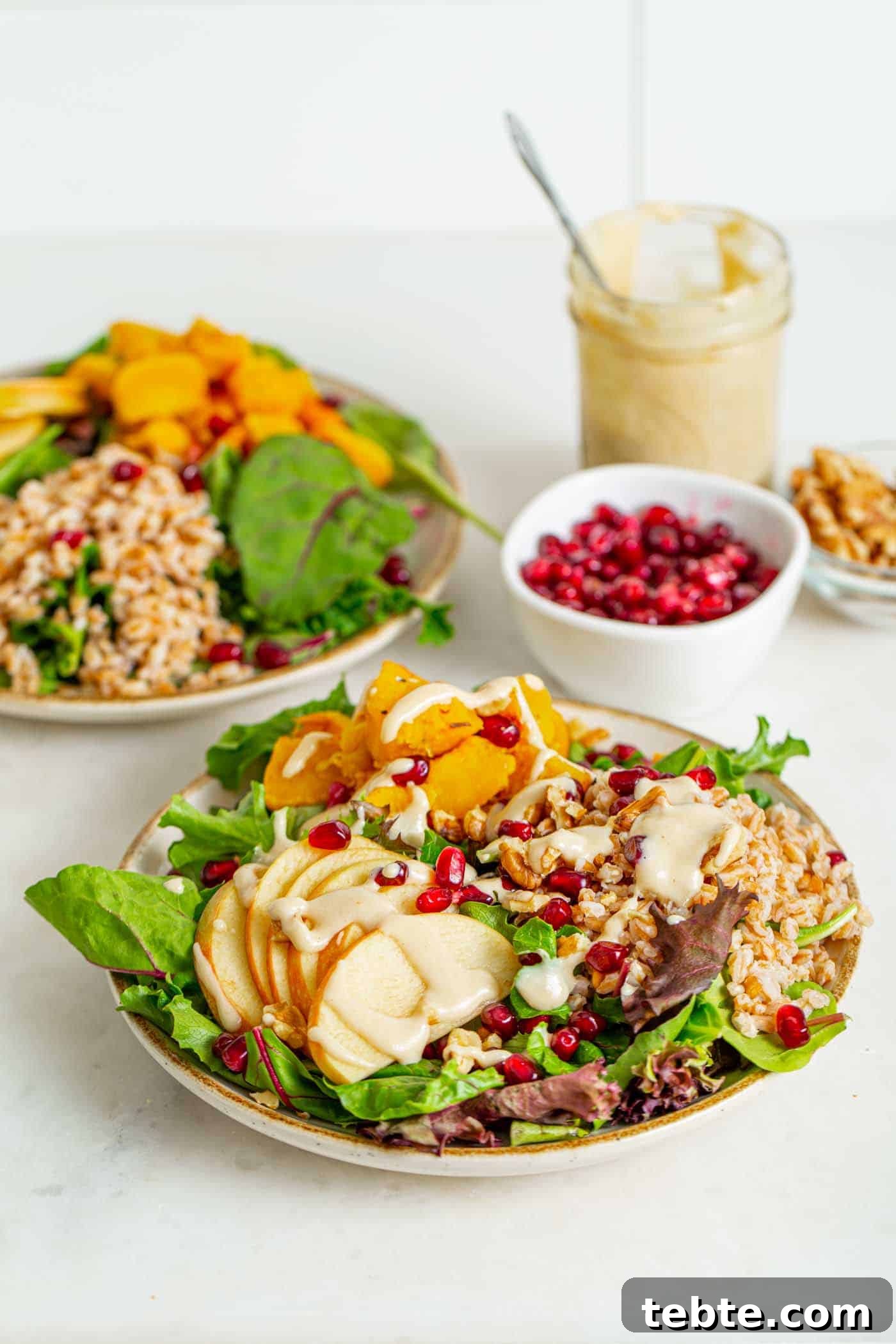 41 Fresh Fall Salad Recipes to Try This Season 22 One up close shot and one far back shot of bowls of salad, with leafy greens, sliced apples, squash, farro, topped with pomegranate seeds and creamy dressing dishes of pomegranate seeds and nuts sit in the background with a jar of extra dressing.