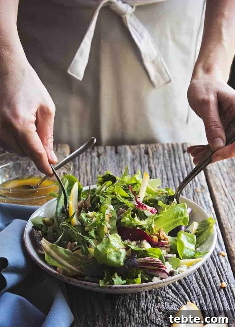 41 Fresh Fall Salad Recipes to Try This Season 36 A person wearing an apron holding two utensils, tossing the salad on a wooden table. Salad is in a white bowl with greens, red lettuce, pears, chunks of cheese, and chopped nuts. Extra dressing if off to the side.