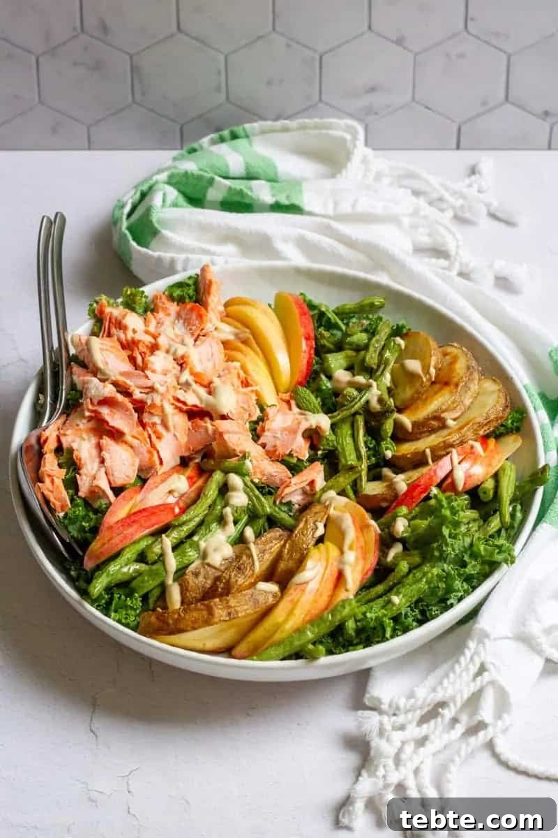41 Fresh Fall Salad Recipes to Try This Season 41 A white bowl sitting on a green striped towel filled with salad ingredients including flaked salmon, sliced potatoes, greens, apples and green beans. The salad is drizzled with a creamy dressing. Serving utensils are sticking out of the bowl.