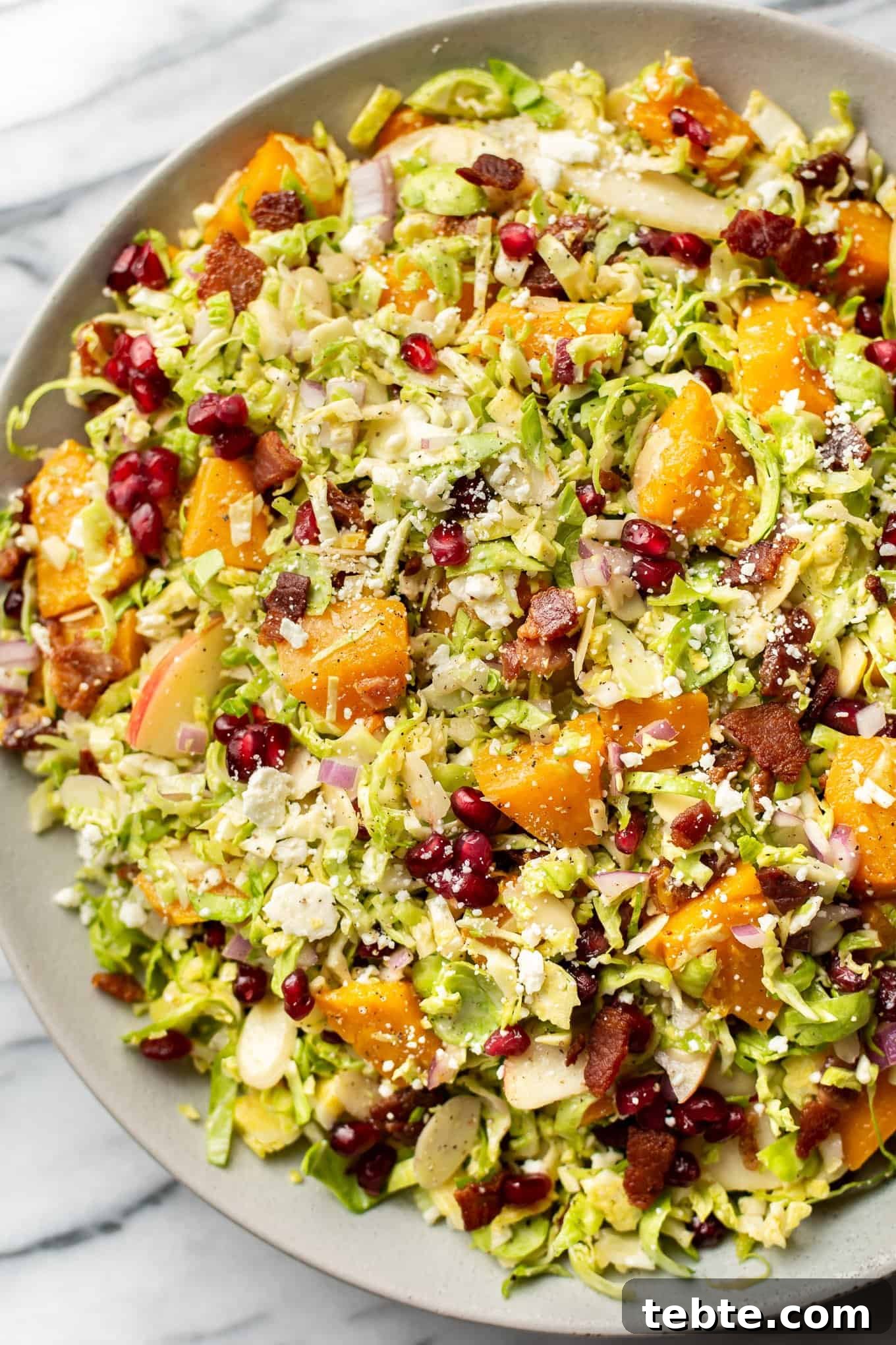 41 Fresh Fall Salad Recipes to Try This Season 43 A close up of the salad sitting in a light colored bowl on a marble surface topped with bacon, pomegranate seeds, shaved brussels sprouts, diced squash, and cheese crumbles.