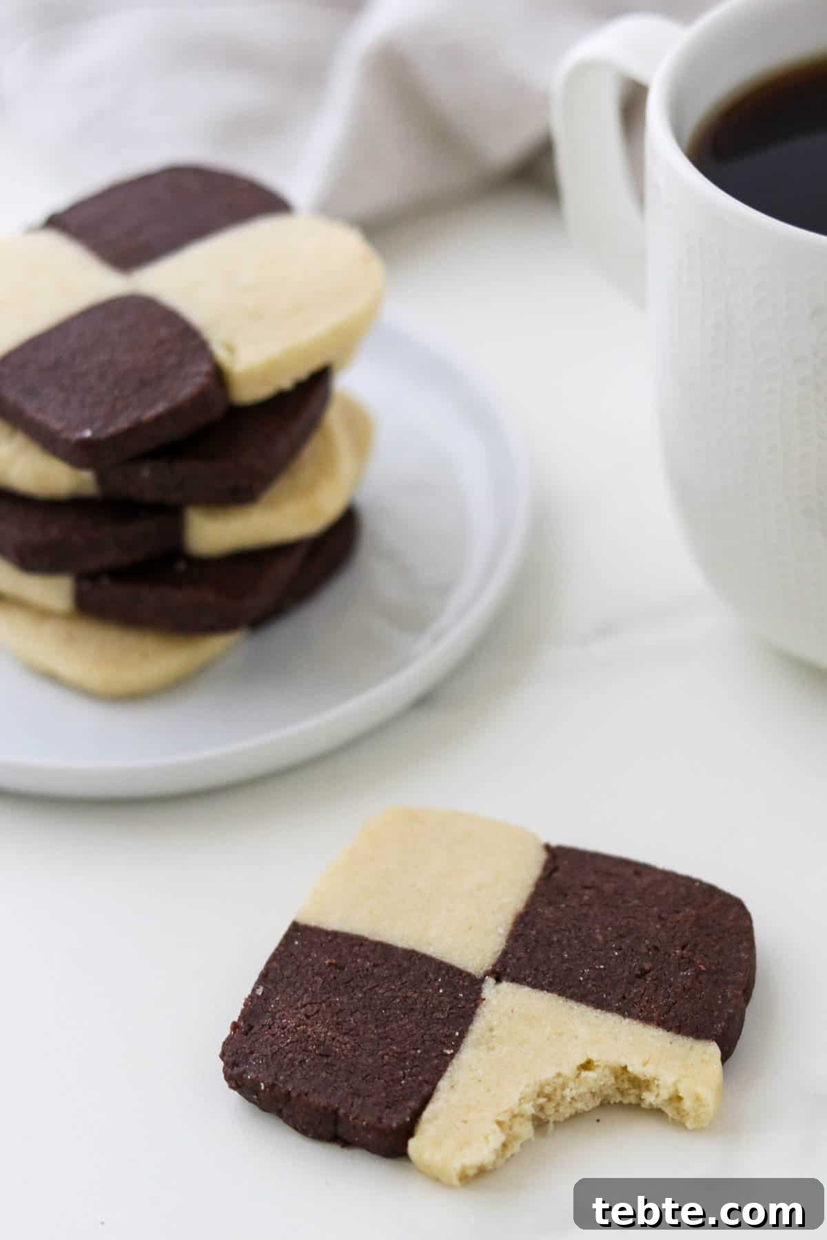 Checkerboard cookie with a bite out of it next to a stack of cookies and coffee.