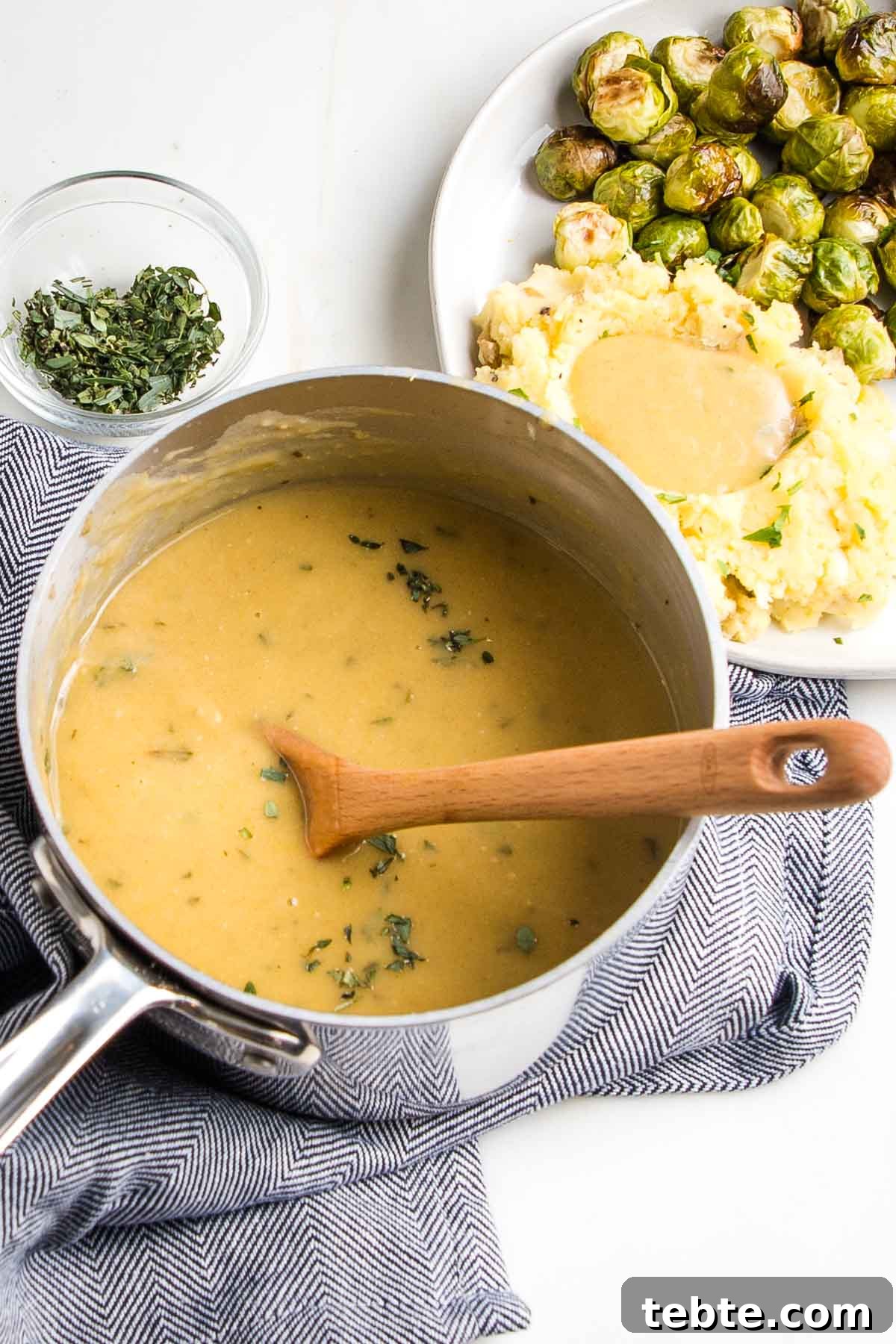 Creamy Vegan Mushroom Gravy Recipe 2 Small saucepan filled with golden gravy, a wooden spoon stirring, next to a plate of mashed potatoes and gravy with brussels sprouts.