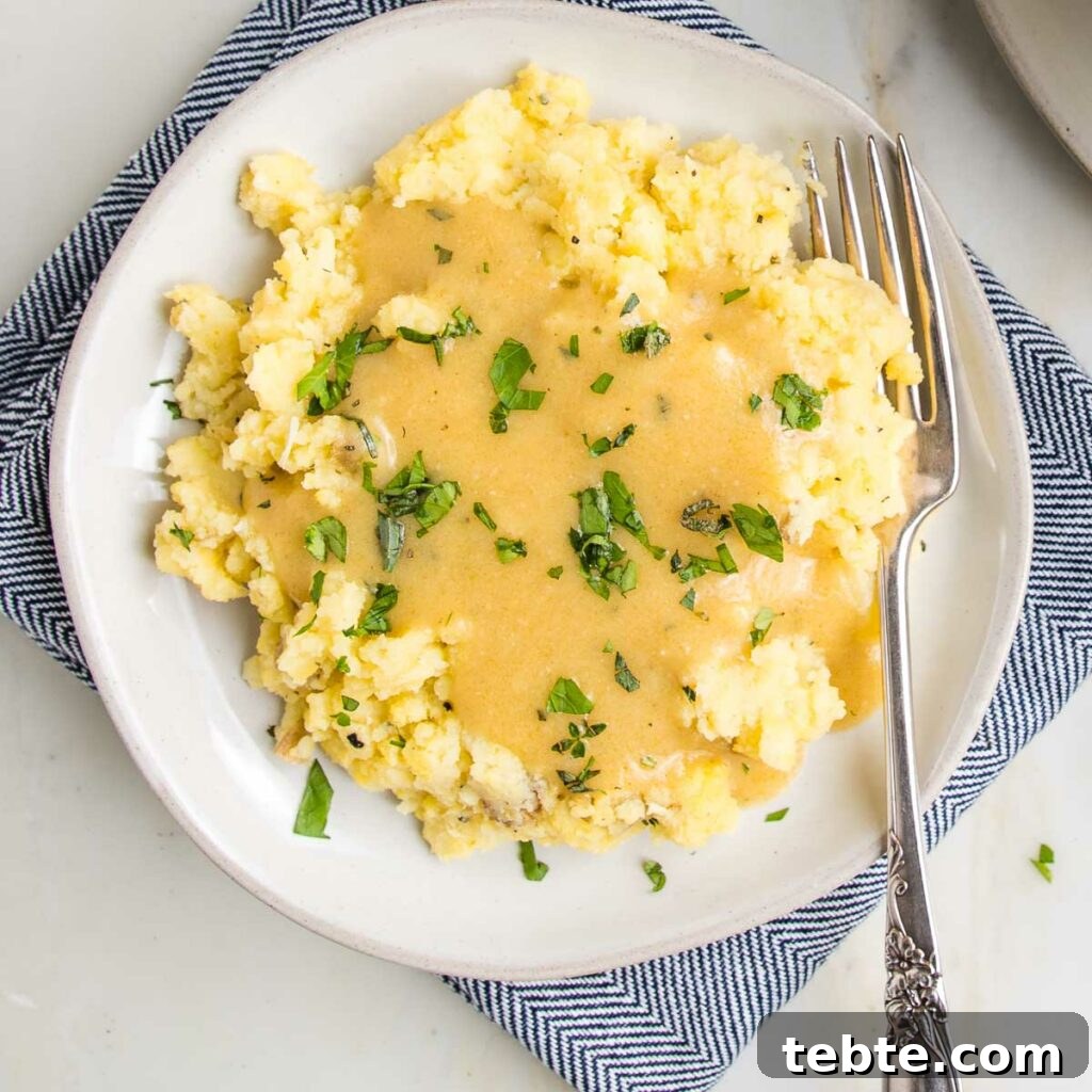 Creamy Vegan Mushroom Gravy Recipe 11 White plate filled with mashed potatoes, smothered in golden vegan gravy with fresh herbs sprinkled on top.