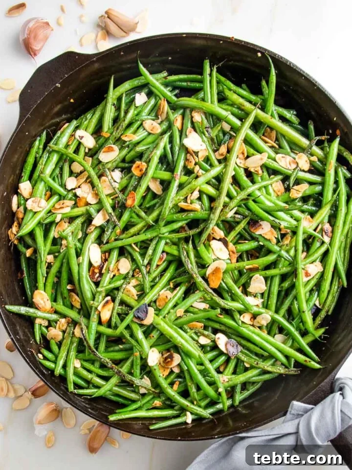 Cast iron pan filled with green beans almondine. There are almond slices, cloves of garlic and crushed red pepper scattered around the pan and a grey napkin tied around the cast iron handle.