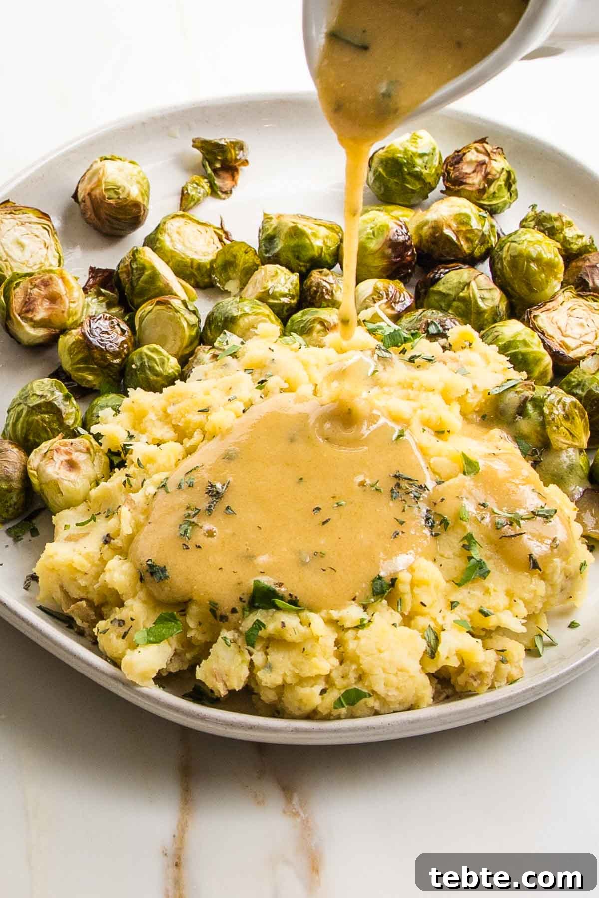 Creamy Vegan Mushroom Gravy Recipe 10 White pitcher pouring vegan gravy over a plate of mashed potatoes and brussels sprouts