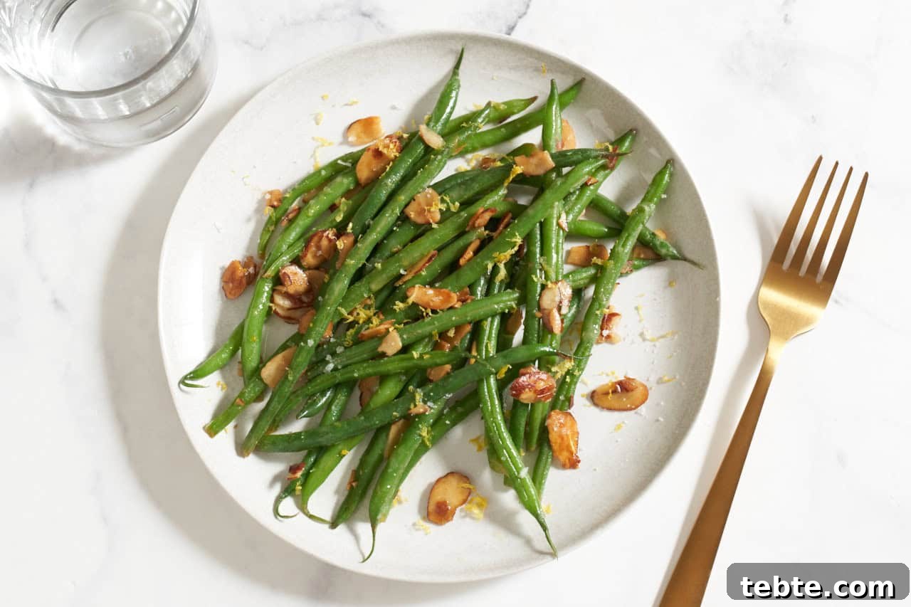 Green beans amandine on a white plate, a gold fork is to the right and a glass of water is on the left.