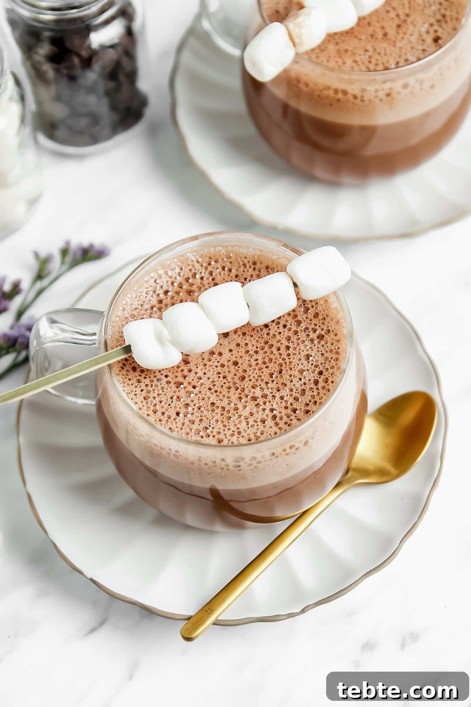 vegan hot chocolate in a clear glass with a stick of marshmallows on top of them. more hot chocolate in the background