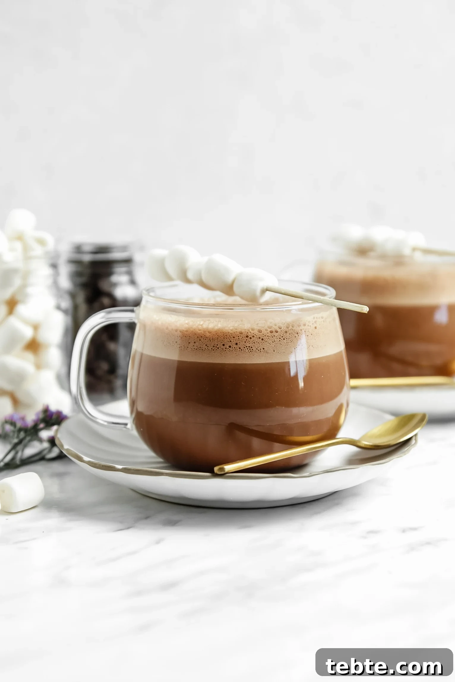 vegan hot chocolate in a clear glass with a stick of marshmallows on top of them. more hot chocolate in the background