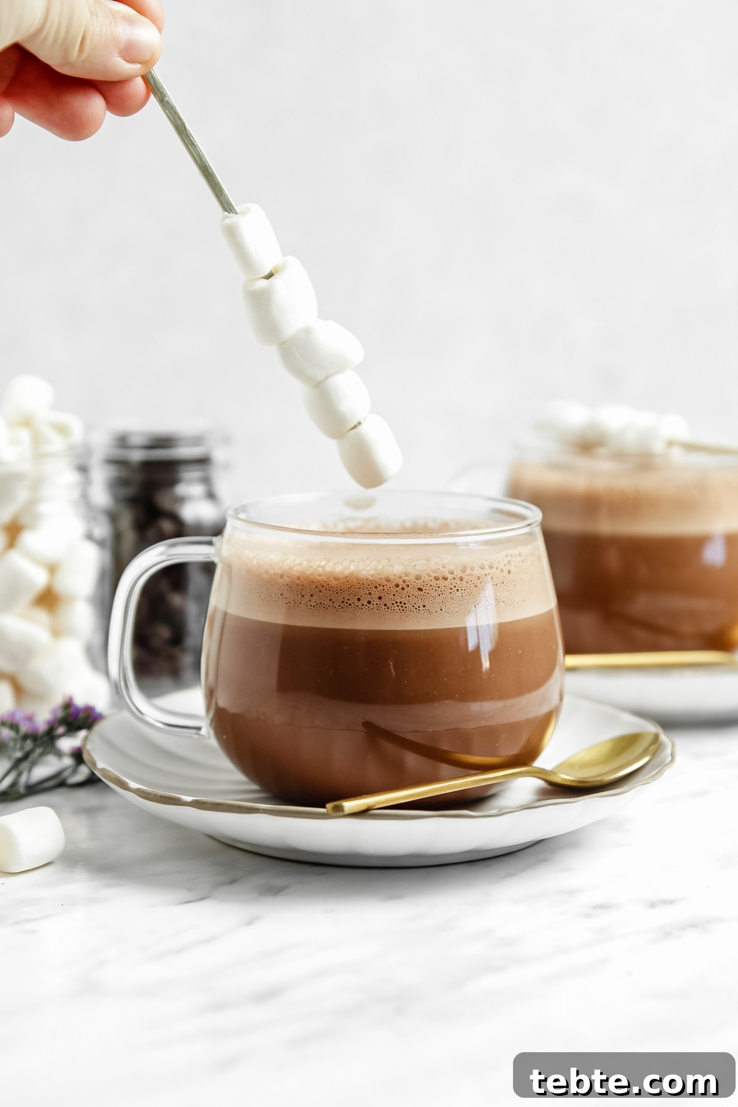 vegan hot chocolate in a clear glass with a stick of marshmallows on top of them. more hot chocolate in the background